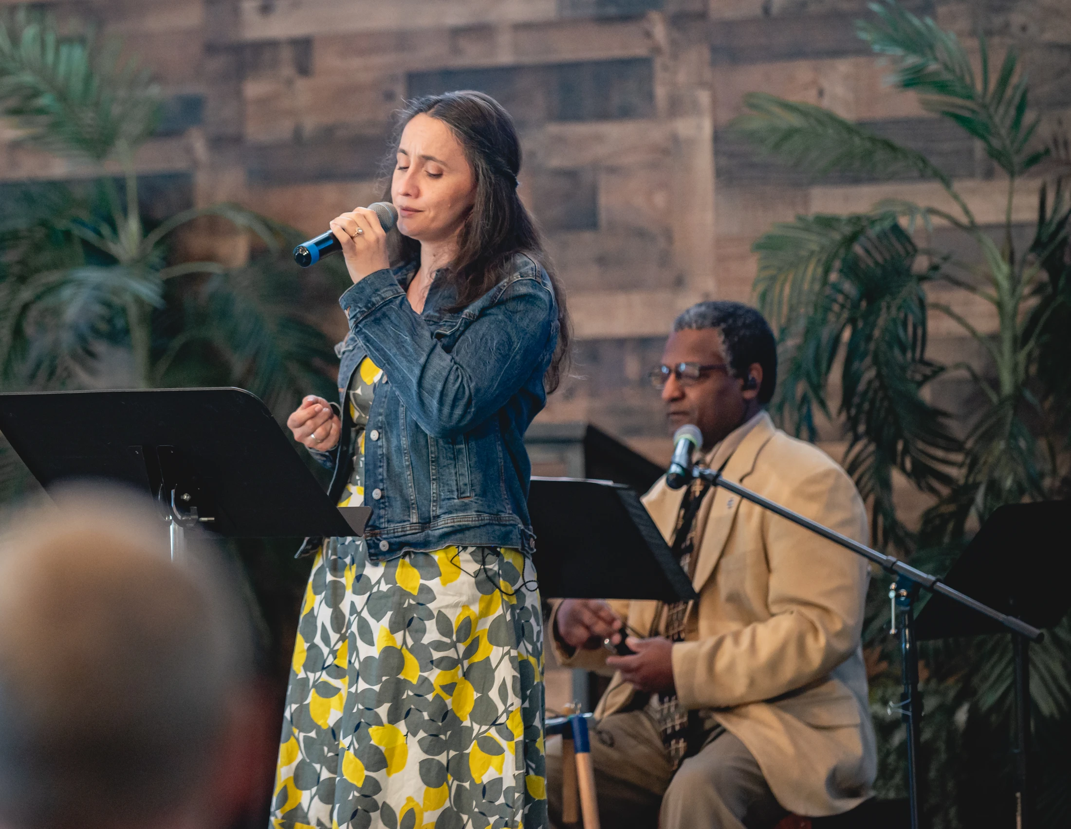 A woman with her eyes closed sings into a microphone on a stage. In the background, a man sits behind her, and the stage is decorated with plants.