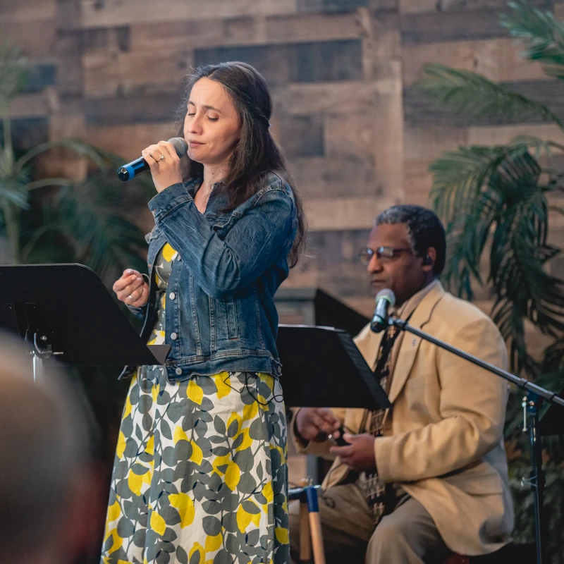 A woman with her eyes closed sings into a microphone on a stage. In the background, a man sits behind her, and the stage is decorated with plants.