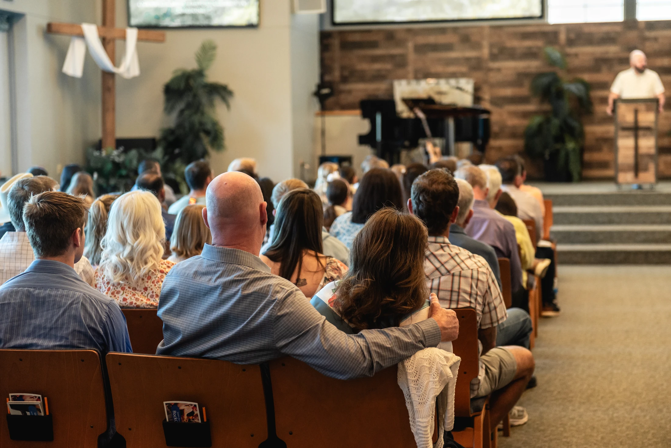 A view from behind a congregation sitting in wooden pews, listening to a pastor give a sermon from a stage. A man has his arm around the woman next to him.