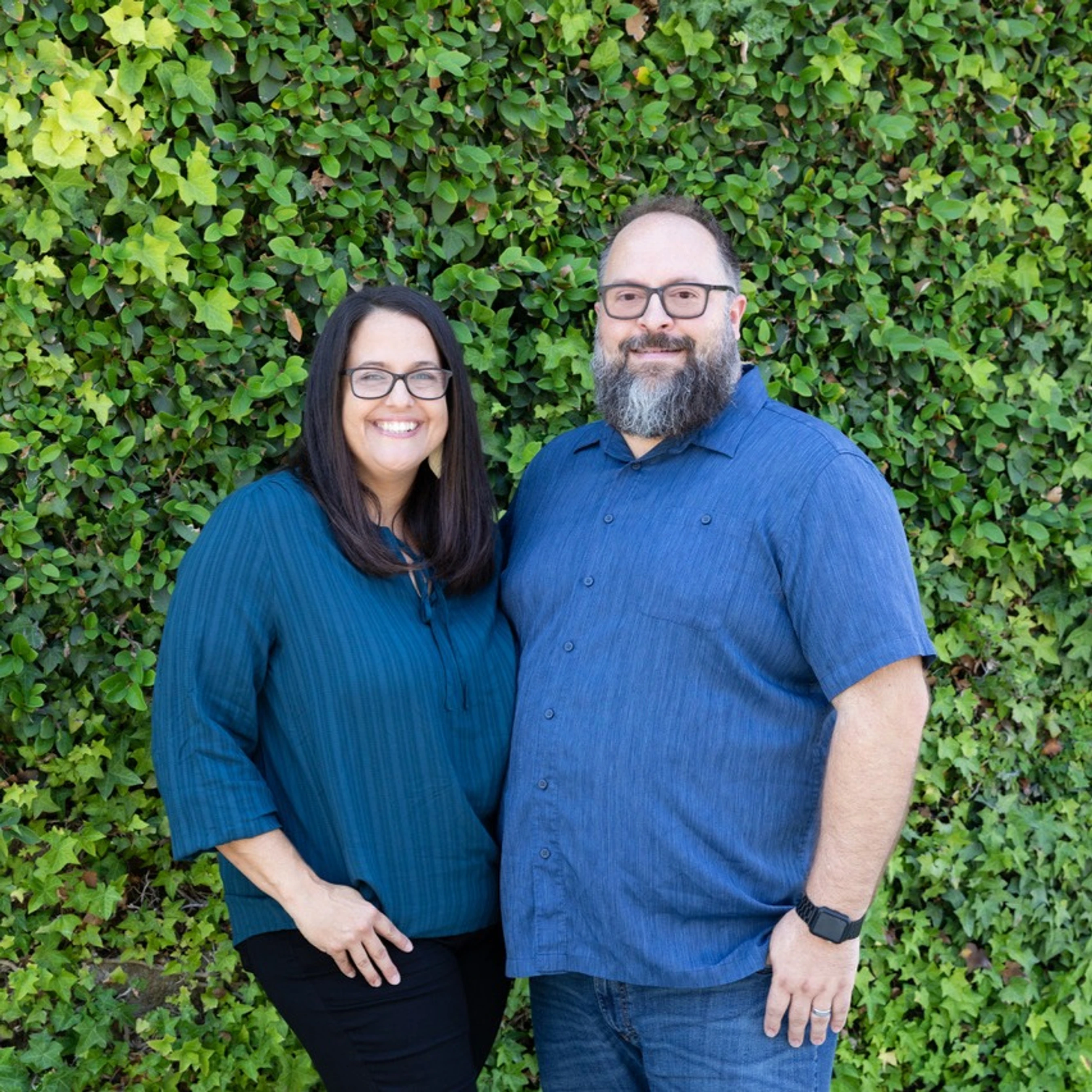 A man with a beard and a woman with long dark hair, both wearing glasses, stand smiling side-by-side in front of a wall completely covered in green ivy.