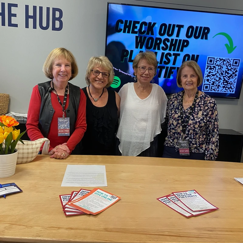 Four smiling women stand behind an information desk labeled 'THE HUB'. A screen behind them advertises a worship playlist with a QR code.