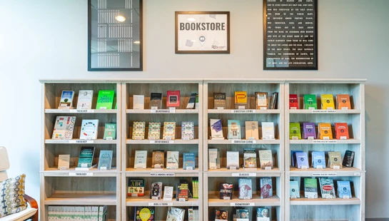 A wide shot of a wooden bookshelf in a bookstore, filled with Christian literature organized by category. A "BOOKSTORE" sign hangs on the wall above.