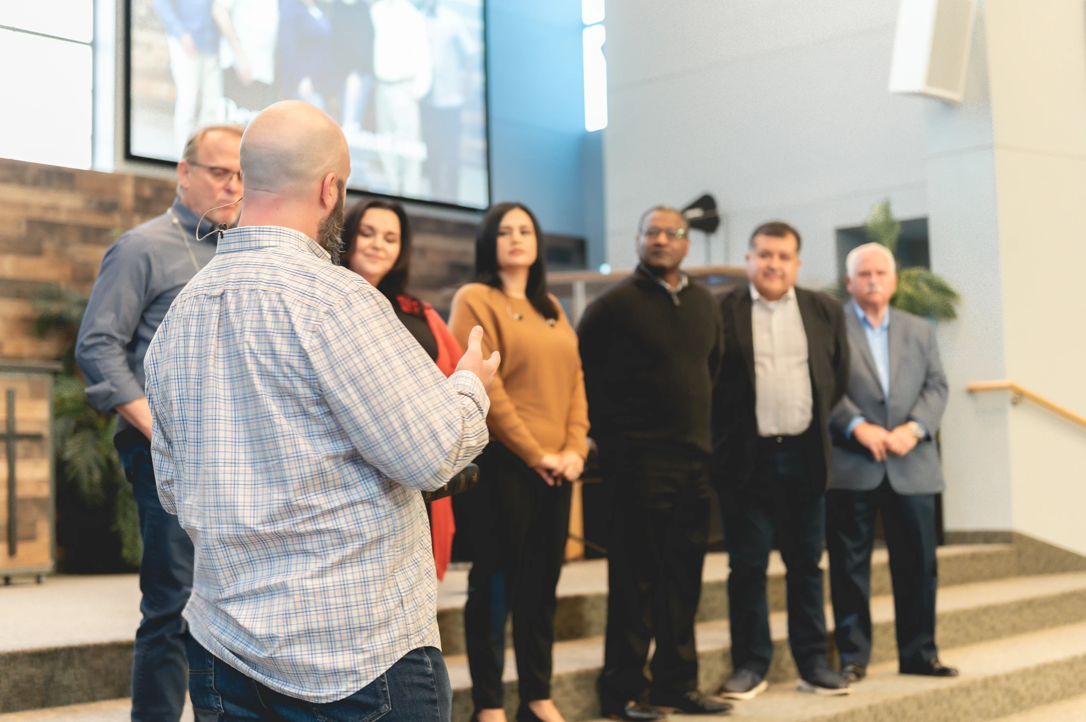 A bald man in a plaid shirt stands with his back to the camera, speaking to a diverse group of people standing on the steps of a church stage.