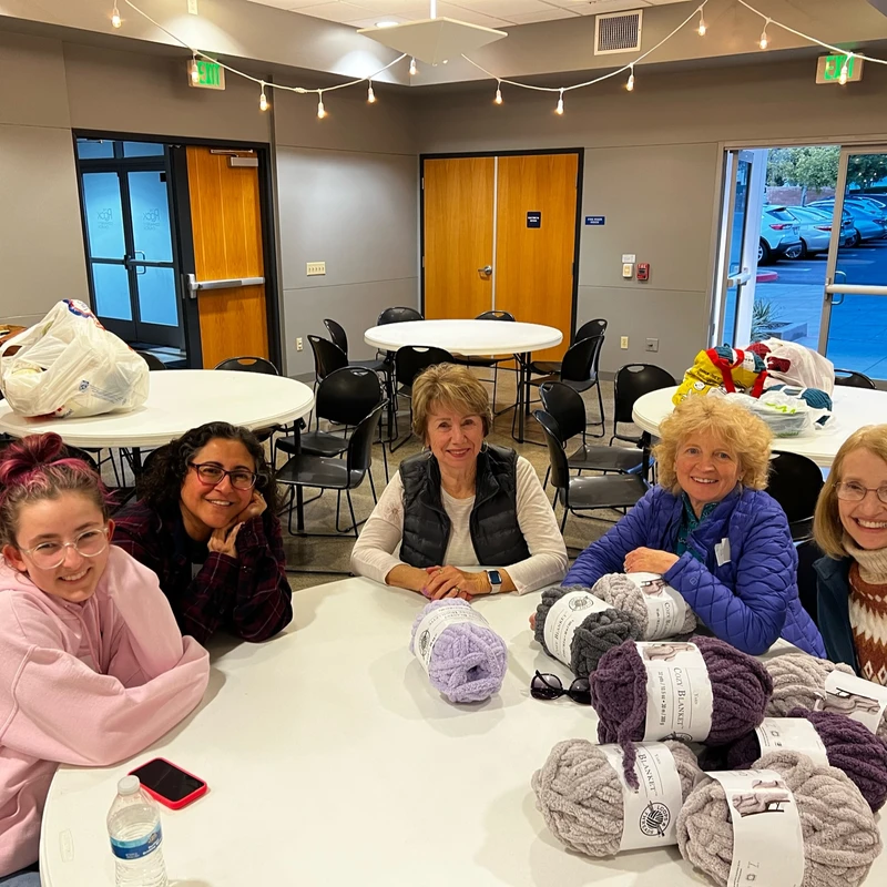 Five women of varying ages sit smiling around a white table covered with skeins of chunky yarn, ready for a group crafting event in a community room.