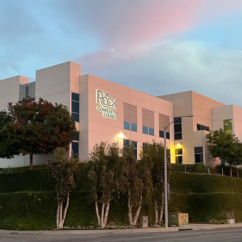 A large, modern, off-white building for The Rock Community Church, with its sign illuminated against a pink and blue sky at dusk.