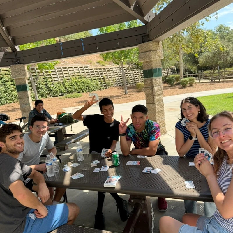 A group of seven smiling young people sit around a picnic table under a pergola, playing a card game on a sunny day in a park.