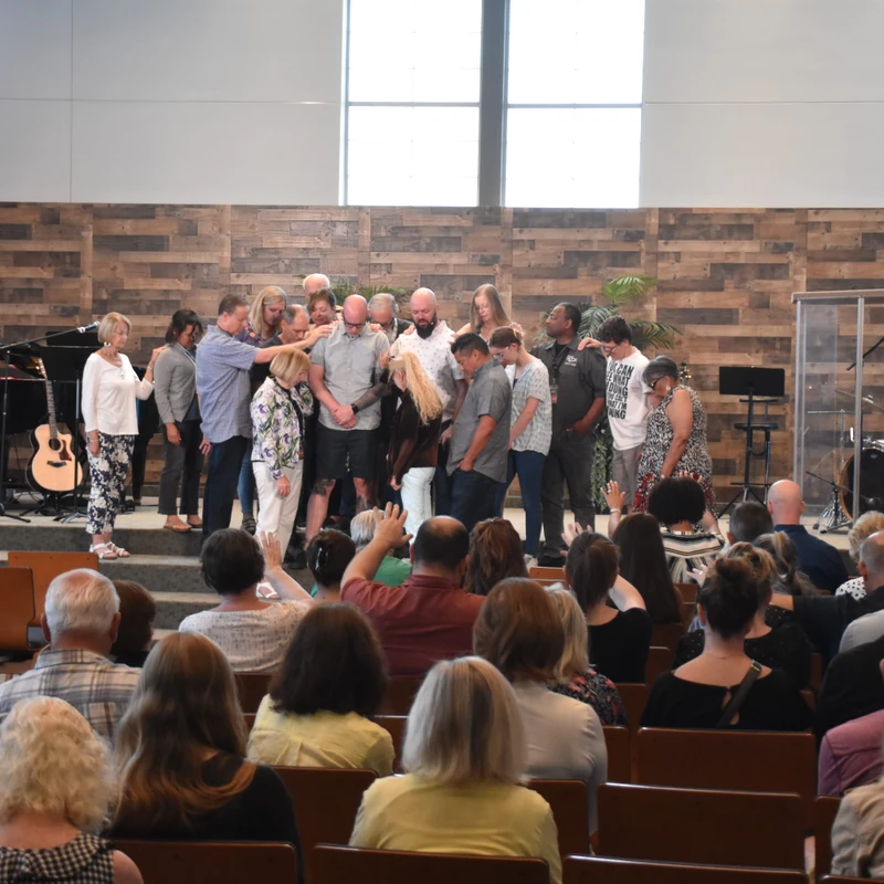 A congregation watches from pews as a group of people stand on stage in a circle, praying over several individuals by laying hands on them.