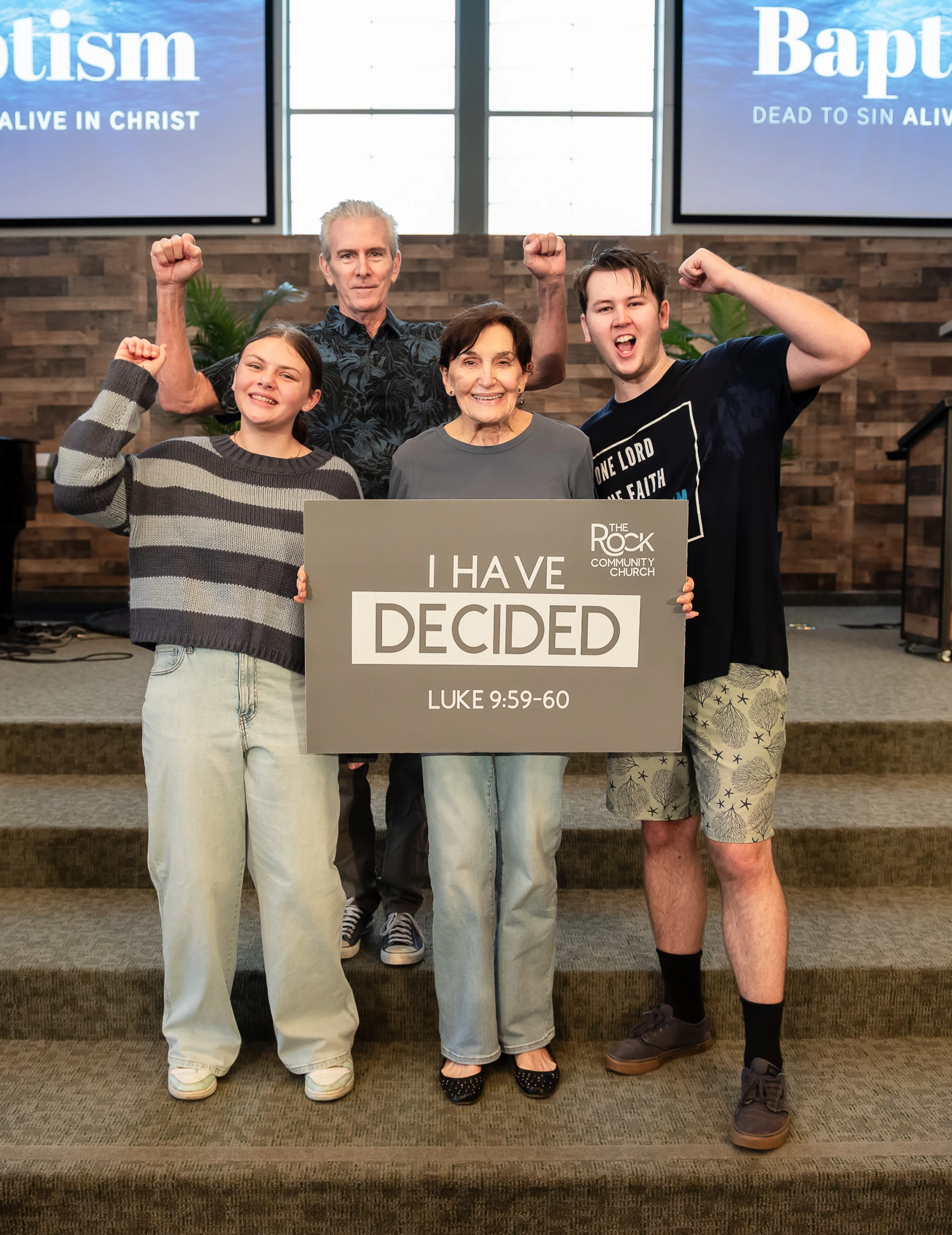 A family of four stands on church steps, smiling and raising their fists. They hold a sign that reads 'I Have Decided' after a baptism ceremony.