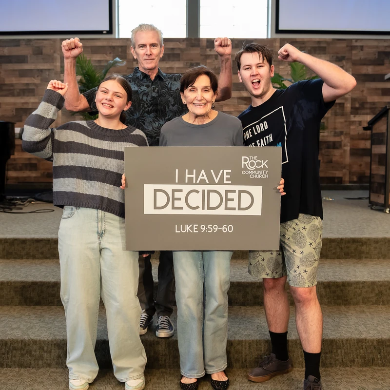 A family of four stands on church steps, smiling and raising their fists. They hold a sign that reads 'I Have Decided' after a baptism ceremony.