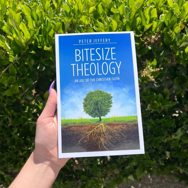 A person's hand with purple nails holds up the book 'Bitesize Theology' by Peter Jeffery in front of a leafy green bush on a sunny day.