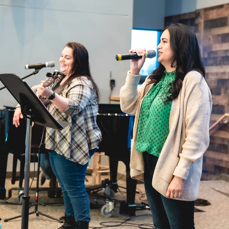 Two women lead music on a stage. One sings into a microphone while the other smiles and plays an acoustic guitar. A piano is visible in the background.