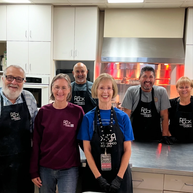 A group of six smiling adult volunteers wearing aprons with 'The Rock Community Church' logo pose for a photo in a commercial kitchen.