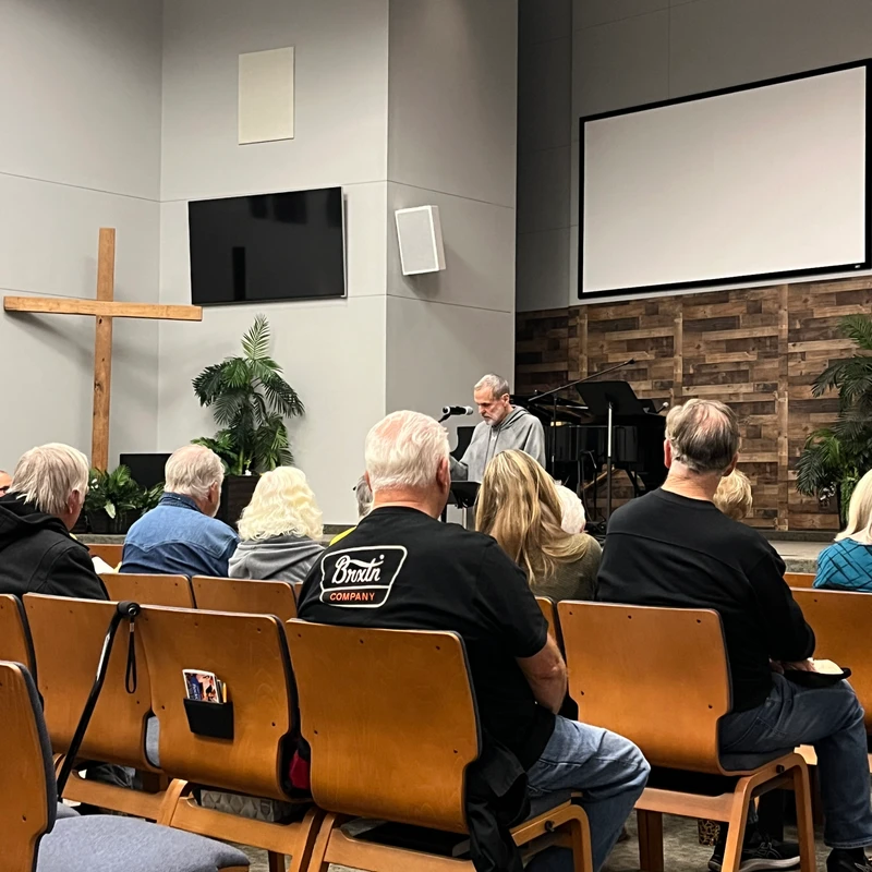 A view from behind an audience sitting in wooden chairs, listening to a man speaking from a stage in a modern church hall with a large cross on the wall.