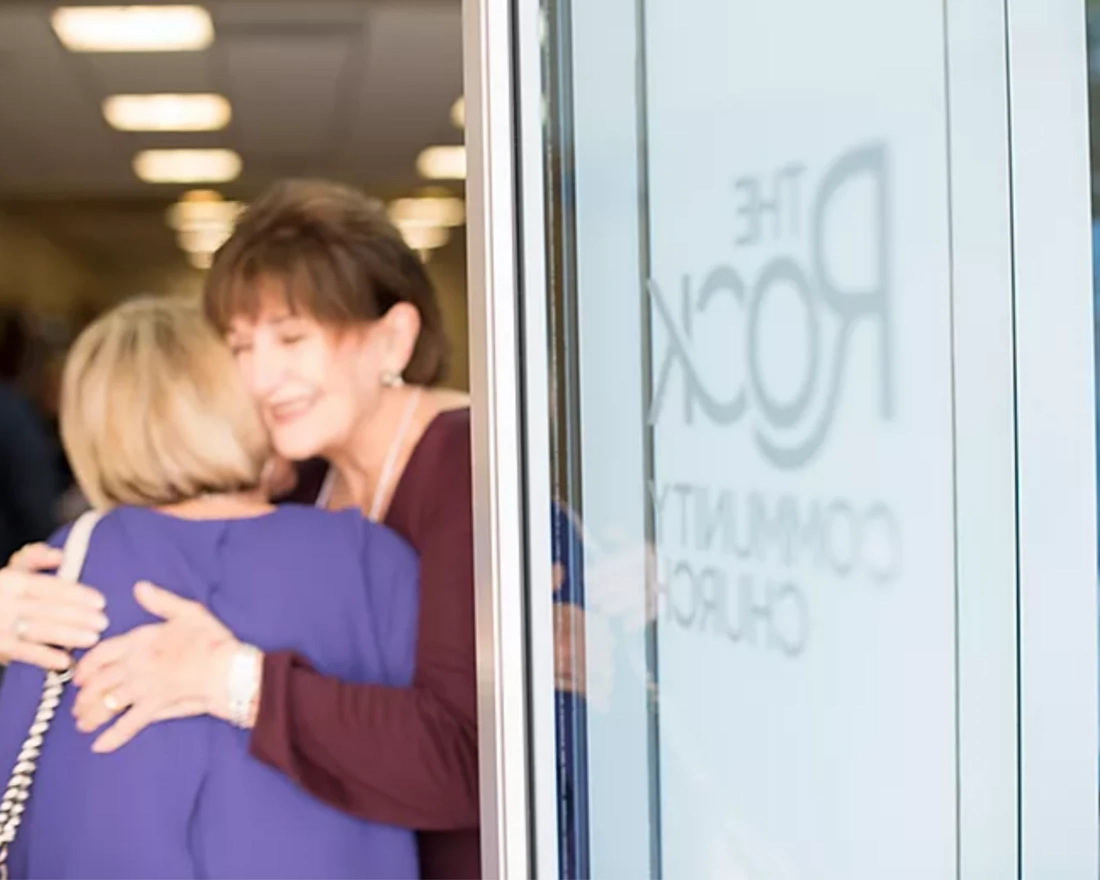 Two women share a warm hug at the entrance of The Rock Community Church, with the church's logo visible on the glass door.