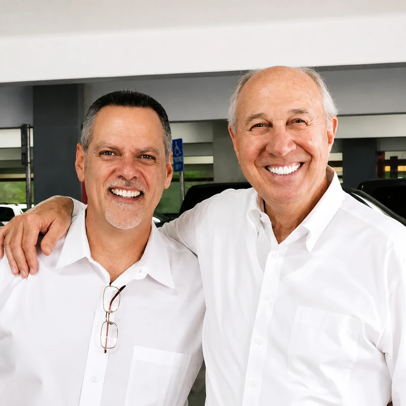 A portrait of two smiling men in white button-down shirts. The older man on the right has his arm around the other's shoulder in a friendly pose.