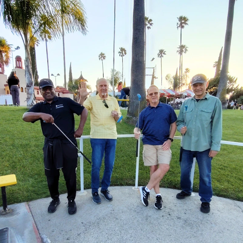 Four men smile for a group photo while holding putters on a miniature golf course surrounded by palm trees at sunset.