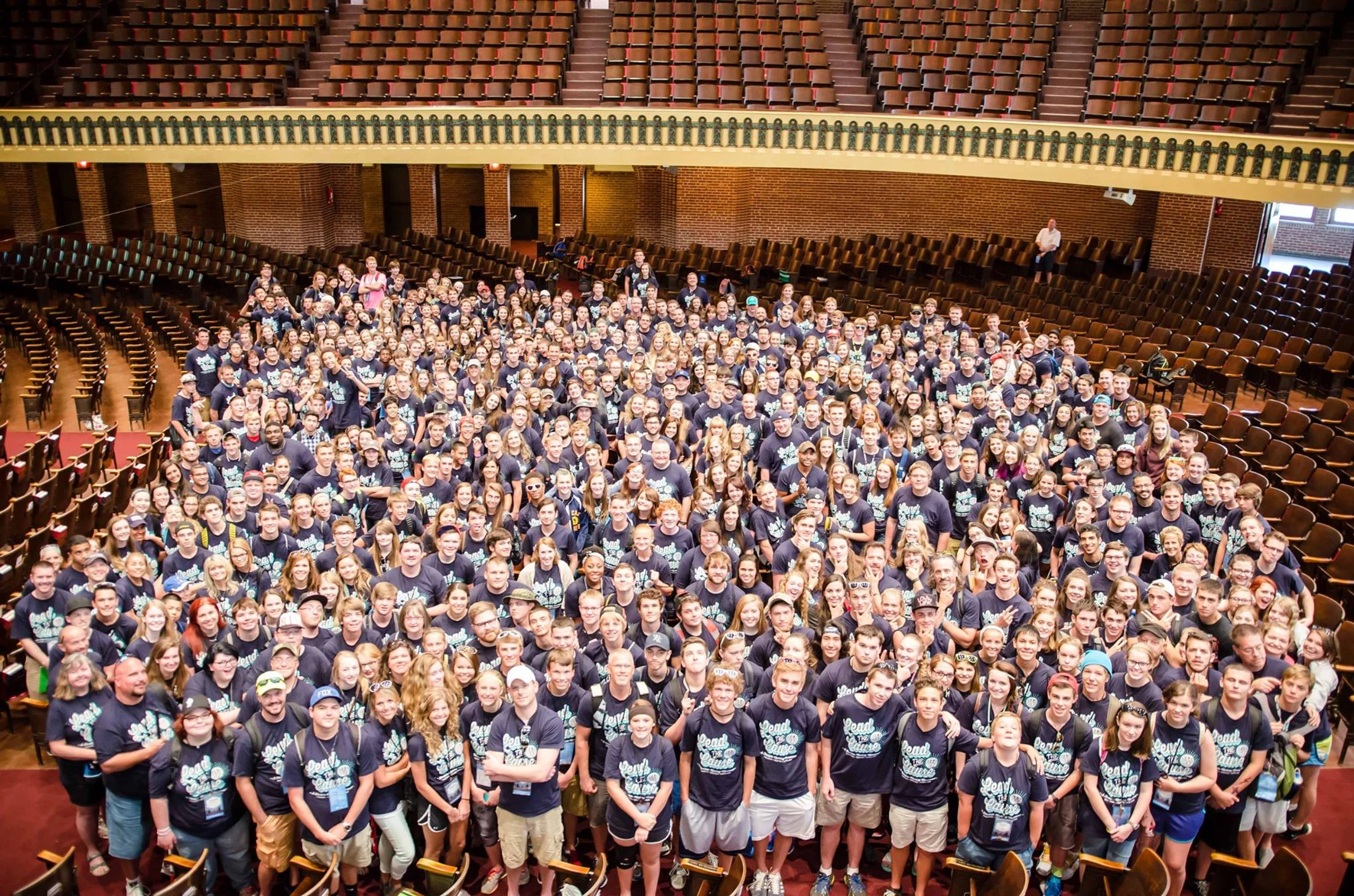 A high-angle photo of a very large group of young people in matching blue t-shirts, smiling and posing for a picture inside a classic-style auditorium.