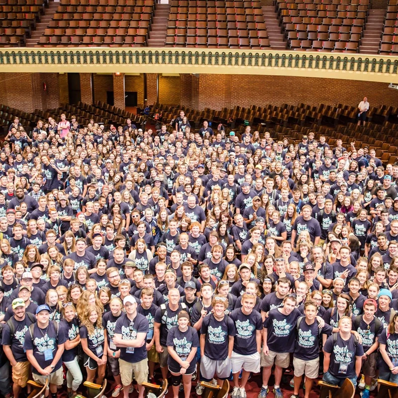 A high-angle photo of a very large group of young people in matching blue t-shirts, smiling and posing for a picture inside a classic-style auditorium.