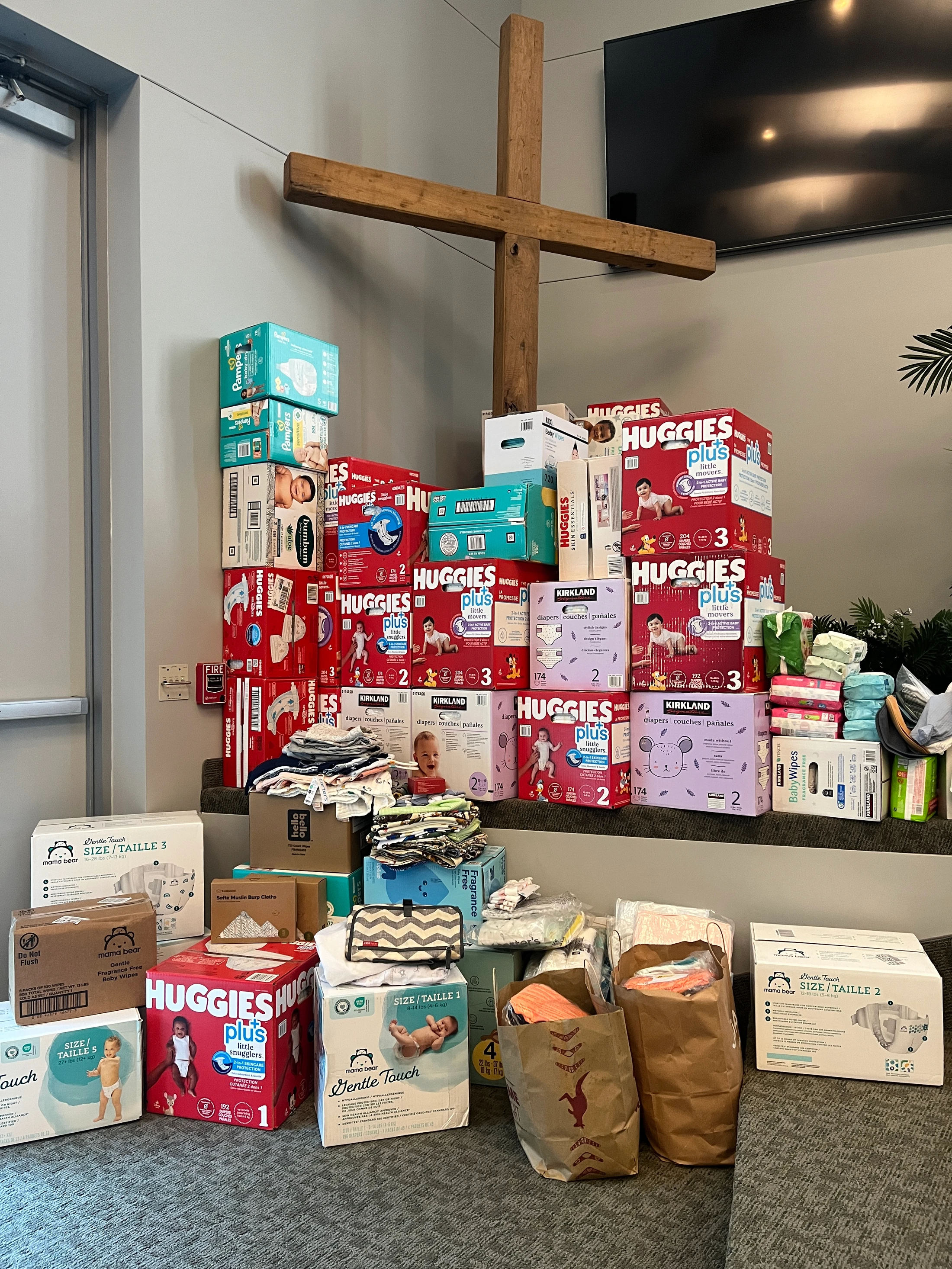 A large pile of donated baby supplies, including many boxes of diapers and baby clothes, stacked in a room in front of a large wooden cross on the wall.