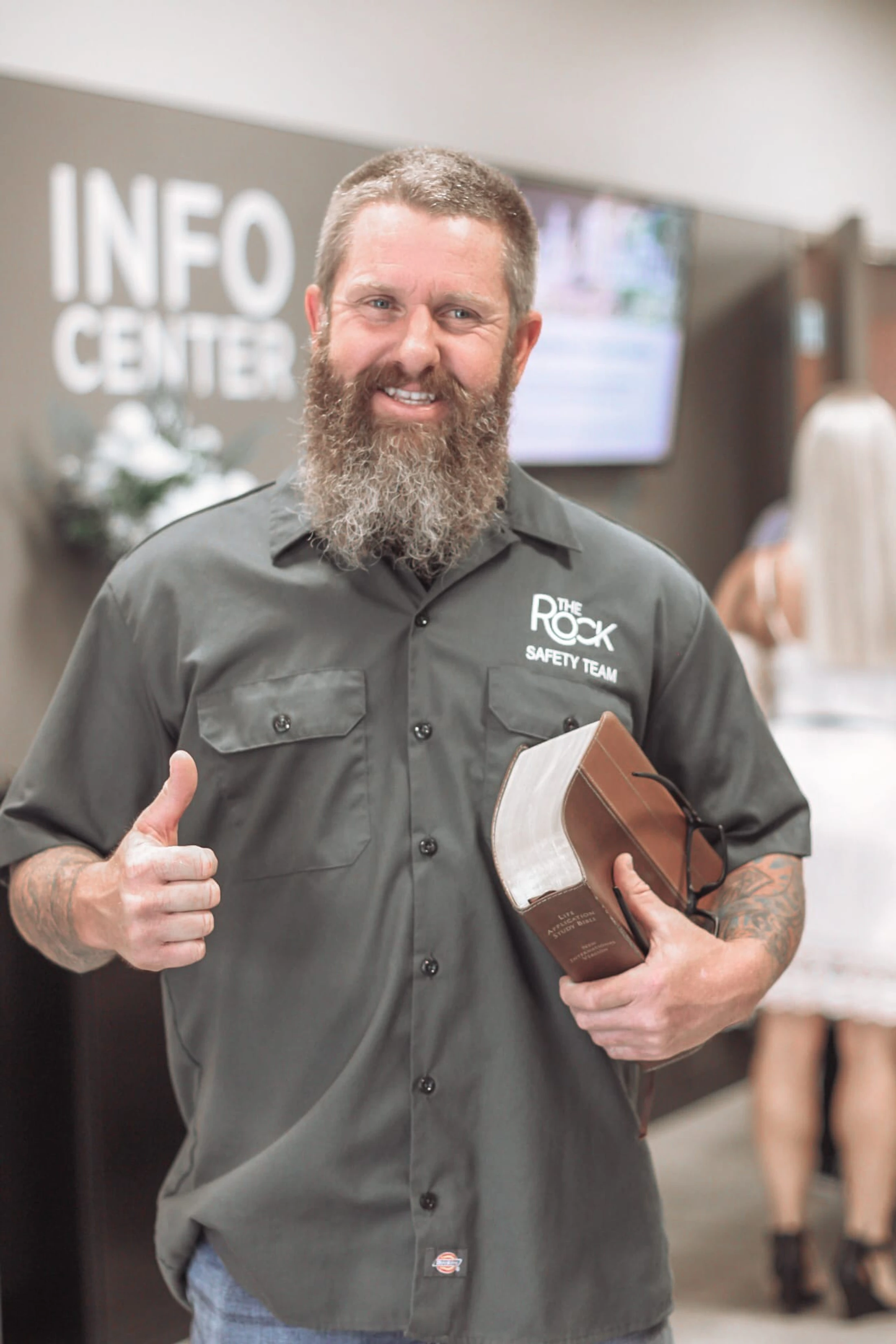 A smiling man with a large beard in a 'The Rock Safety Team' shirt gives a thumbs-up while holding a Bible, standing in front of an Info Center sign.