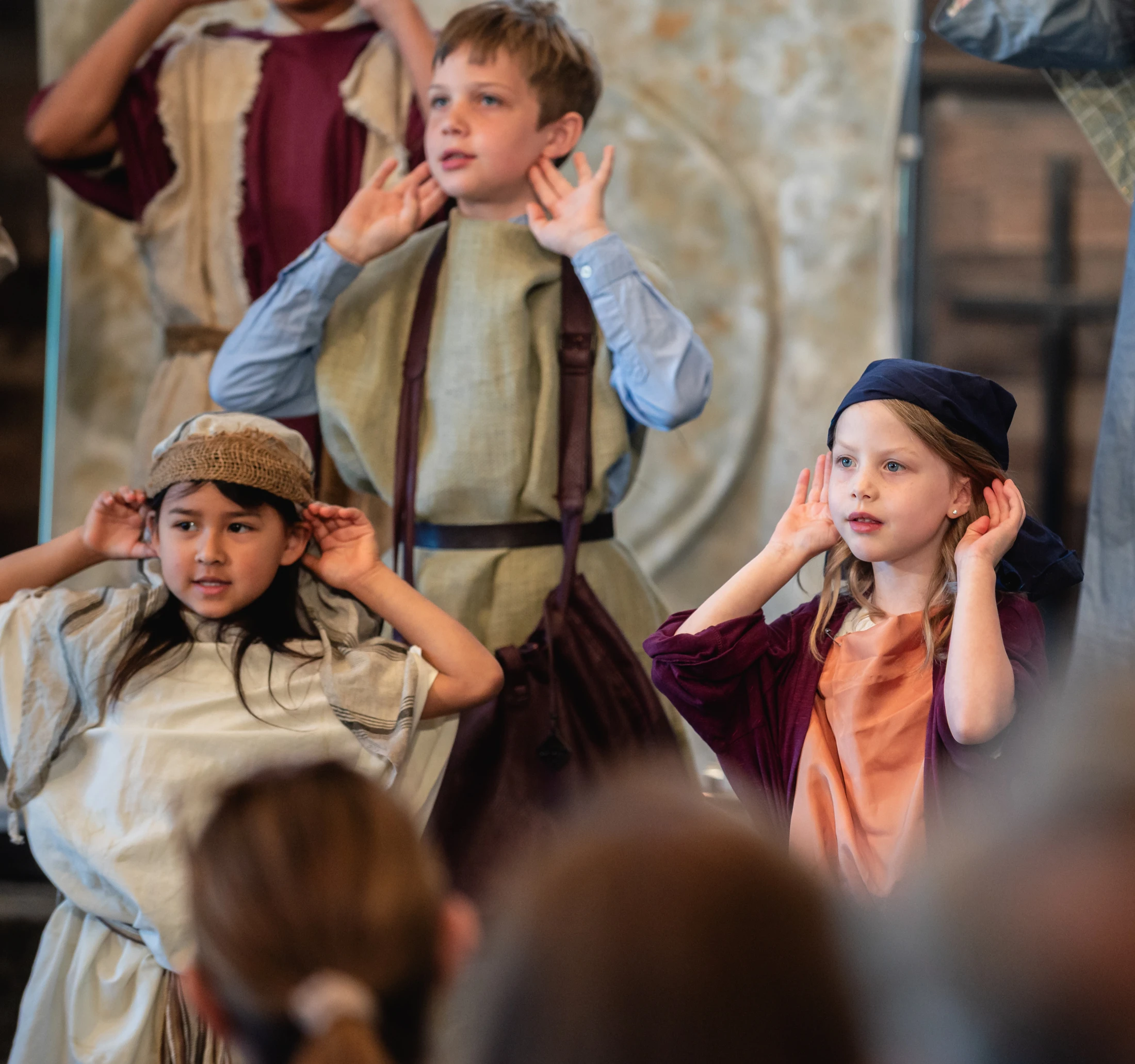 Three children in historical costumes perform on stage. They all have their hands cupped behind their ears as if they are listening intently.
