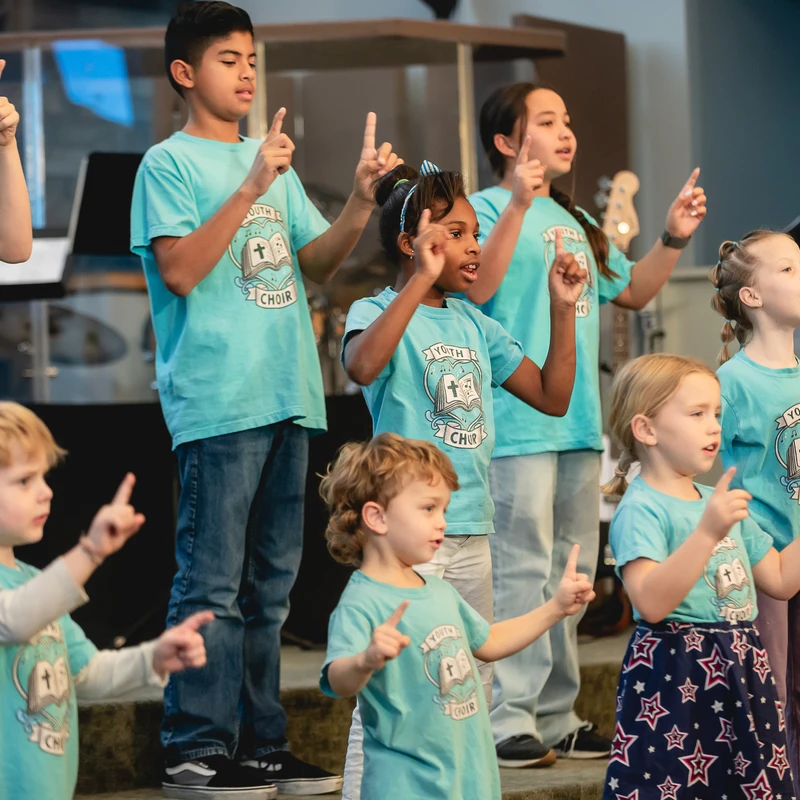 A diverse group of children in matching light blue 'Youth Choir' shirts sing and point their fingers up while performing on a stage.