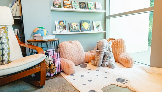 A children's reading corner with a plush pink chair, a stuffed elephant, a paw-print rug, and shelves of books next to a large, bright window.