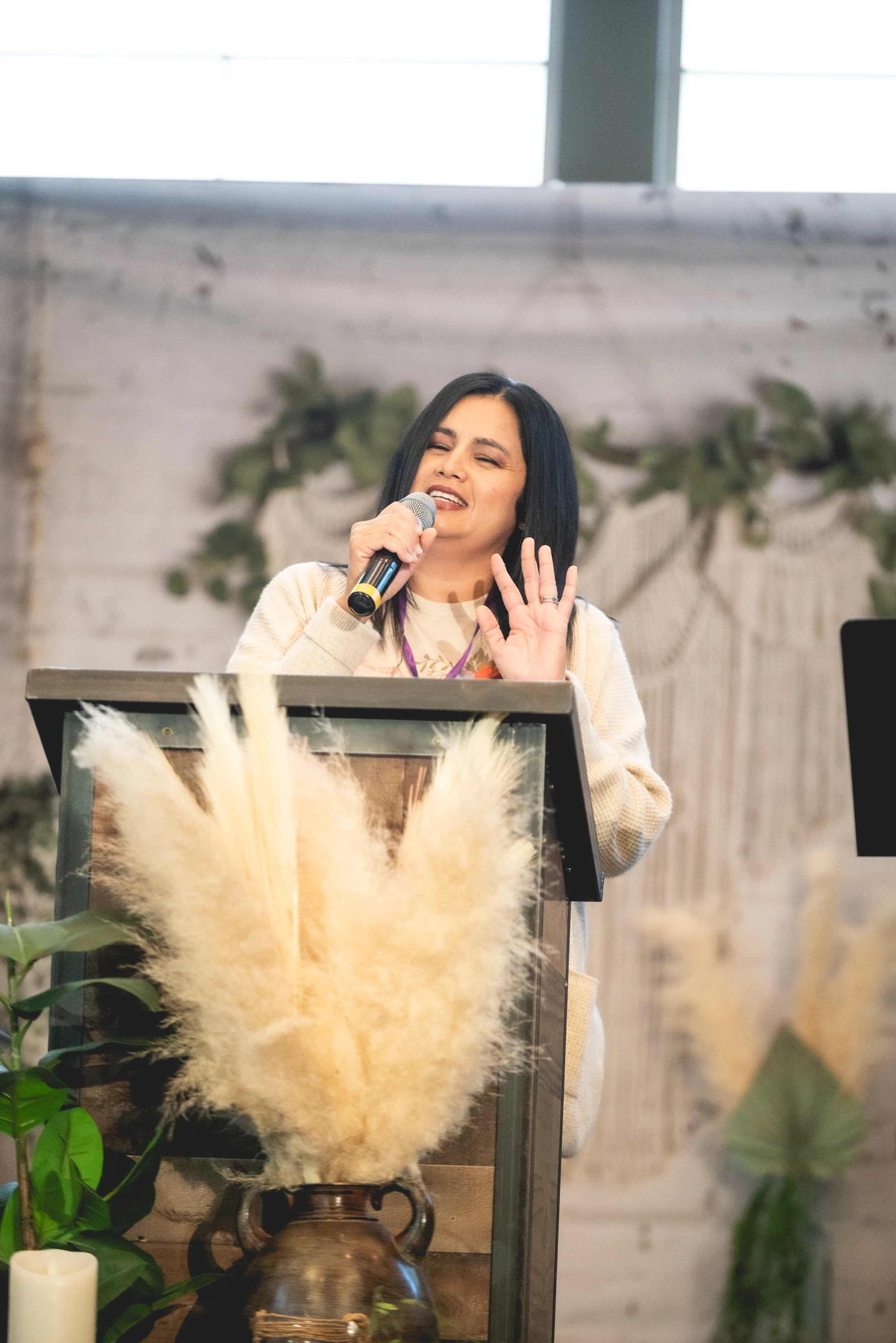 A woman with dark hair stands at a wooden podium decorated with pampas grass, smiling as she speaks into a microphone during an event.