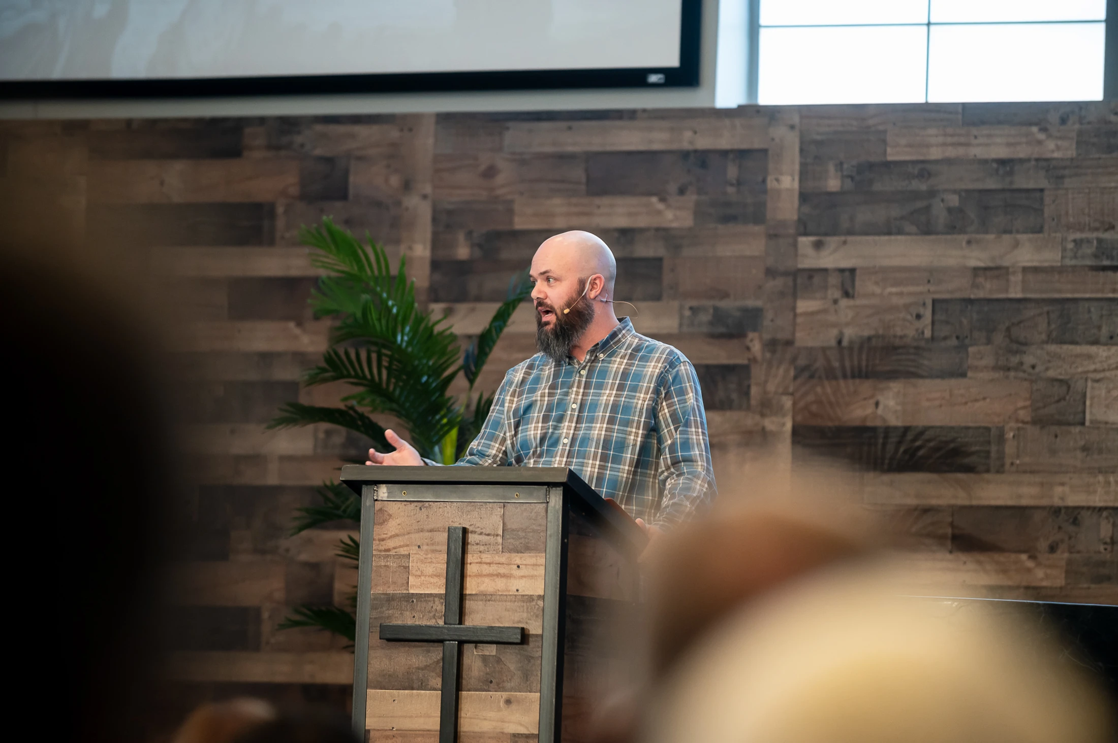 A bald man with a beard wearing a plaid shirt and a lavalier microphone speaks from behind a wooden pulpit with a cross on the front.
