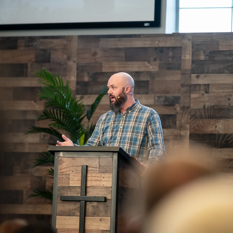 A bald man with a beard wearing a plaid shirt and a lavalier microphone speaks from behind a wooden pulpit with a cross on the front.