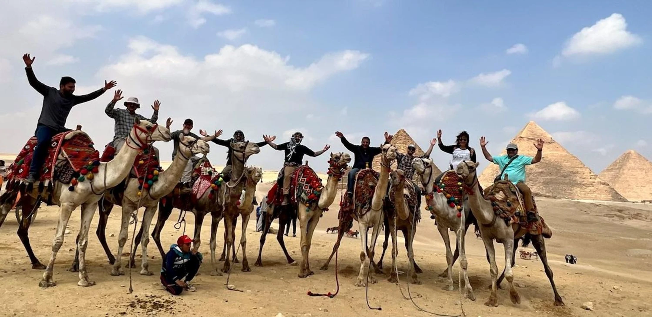 A group of smiling tourists on a line of camels pose for a photo with their arms raised, with the Great Pyramids of Giza in the background.
