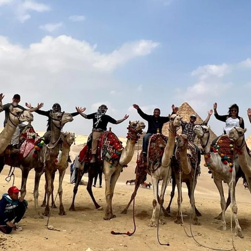 A group of smiling tourists on a line of camels pose for a photo with their arms raised, with the Great Pyramids of Giza in the background.
