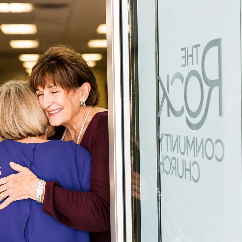 A smiling woman with short brown hair warmly hugs another woman in a doorway next to a glass door with 'The Rock Community Church' logo.