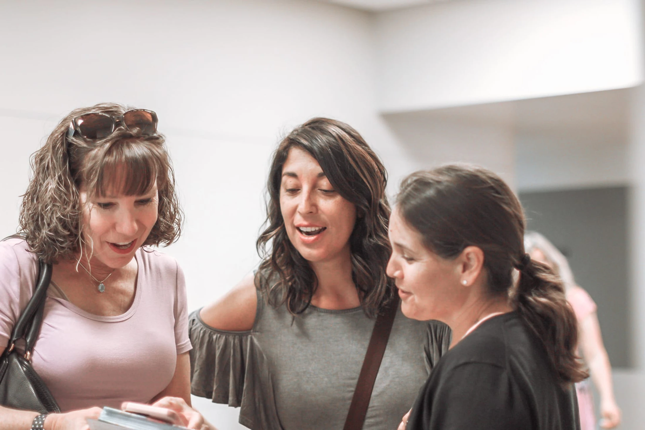 Three women stand close together in a bright room, looking at something one woman holds while the woman in the center speaks.