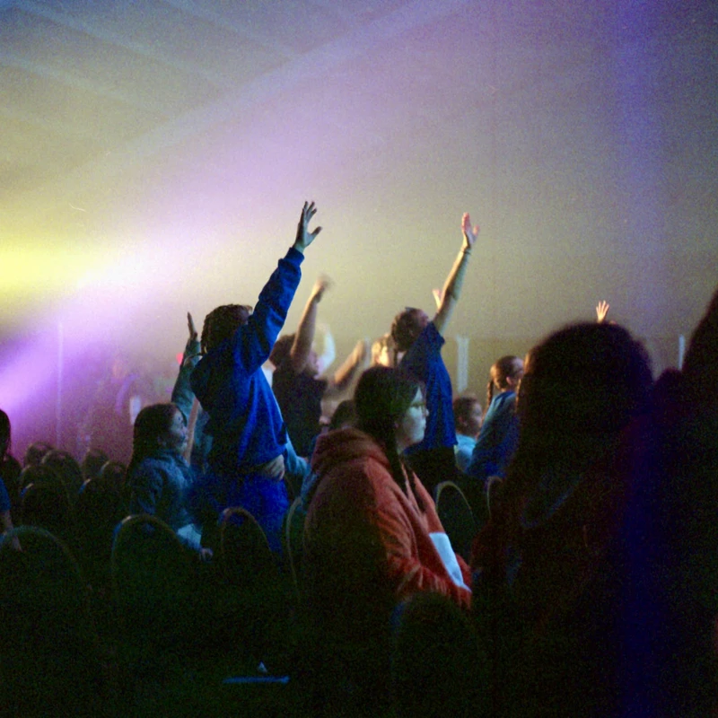 A crowd of people in a dark room sit in chairs, many with their hands raised. Bright yellow and purple stage lights shine down, creating a hazy atmosphere.