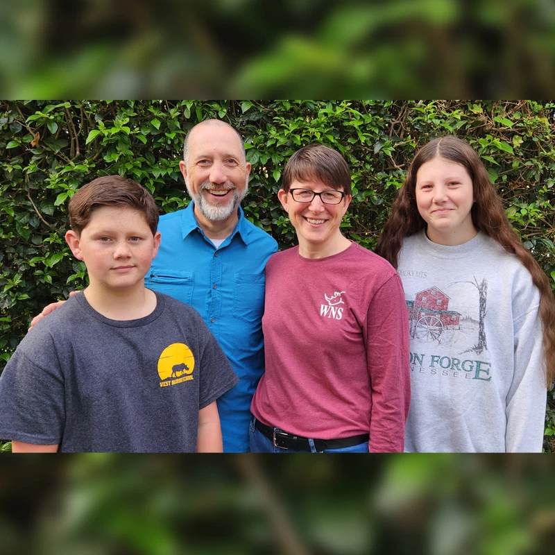 A family of four, a man, a woman, a boy, and a girl, stand close together smiling in front of a wall of green leafy bushes.
