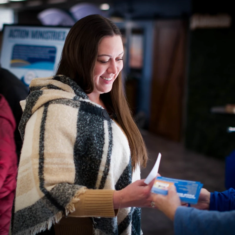 A woman with long brown hair, wearing a plaid poncho, smiles as she accepts a small blue and white pamphlet from another person at an indoor event.