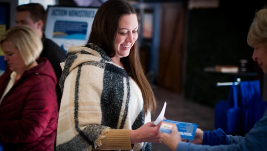 A woman with long brown hair, wearing a plaid poncho, smiles as she accepts a small blue and white pamphlet from another person at an indoor event.