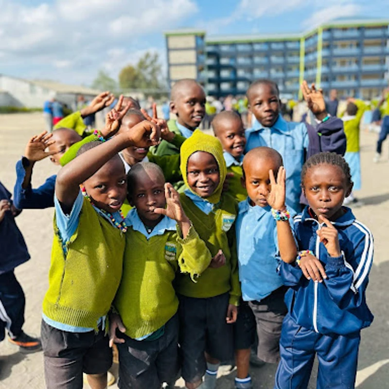 A group of young African schoolchildren in uniforms smiling and posing playfully for the camera in an outdoor schoolyard on a sunny day.