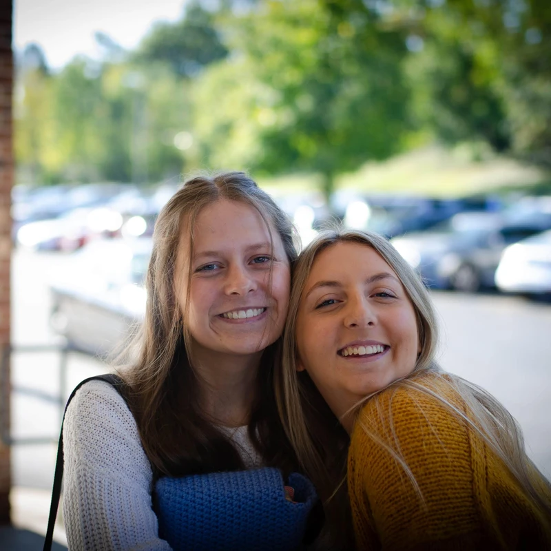 Two young women with blonde and brown hair smile warmly at the camera. They are embracing outdoors with a blurred parking lot and trees in the background.