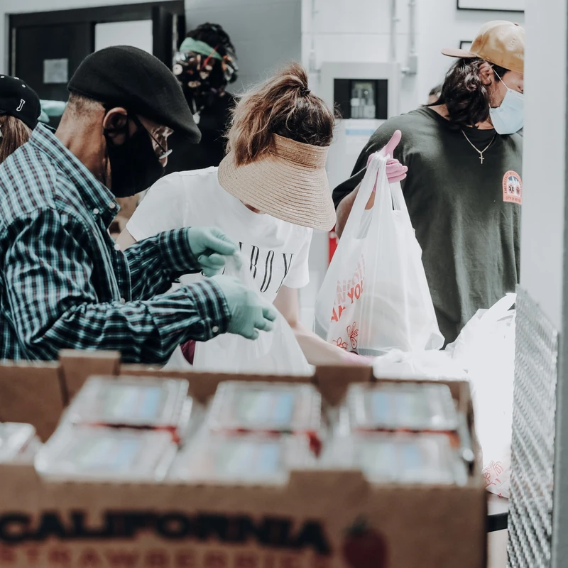 Volunteers at a food bank wearing face masks and gloves pack groceries into bags, with a box of fresh strawberries in the foreground.
