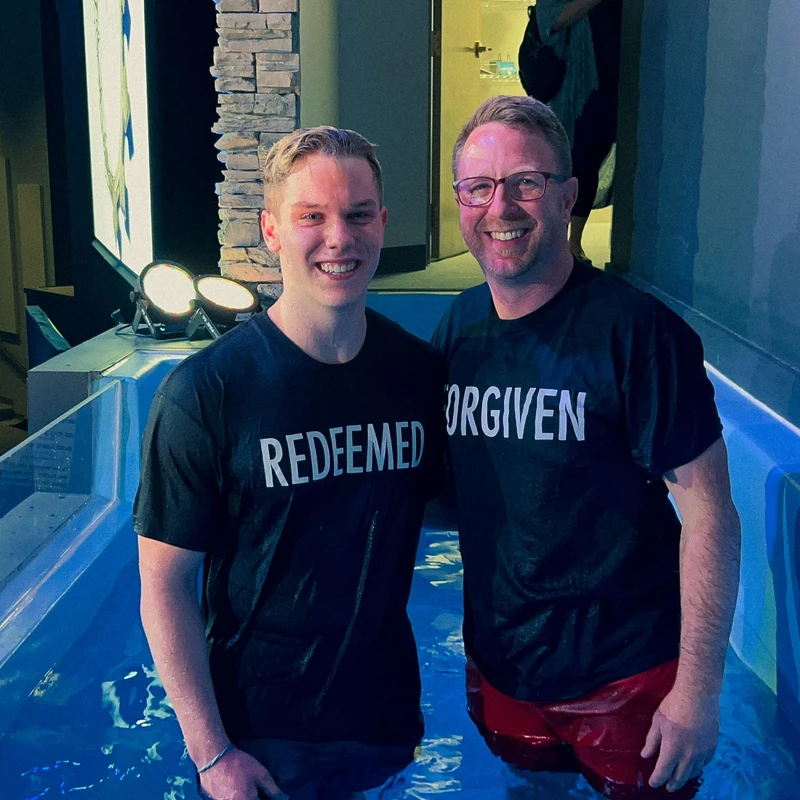 Two smiling men, one younger and one older, stand waist-deep in a baptismal pool. They wear matching black shirts with the words 'REDEEMED' and 'FORGIVEN'.