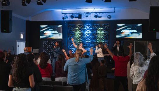 A congregation seen from behind, with many raising their hands in worship toward a band performing on a brightly lit stage in a modern church.