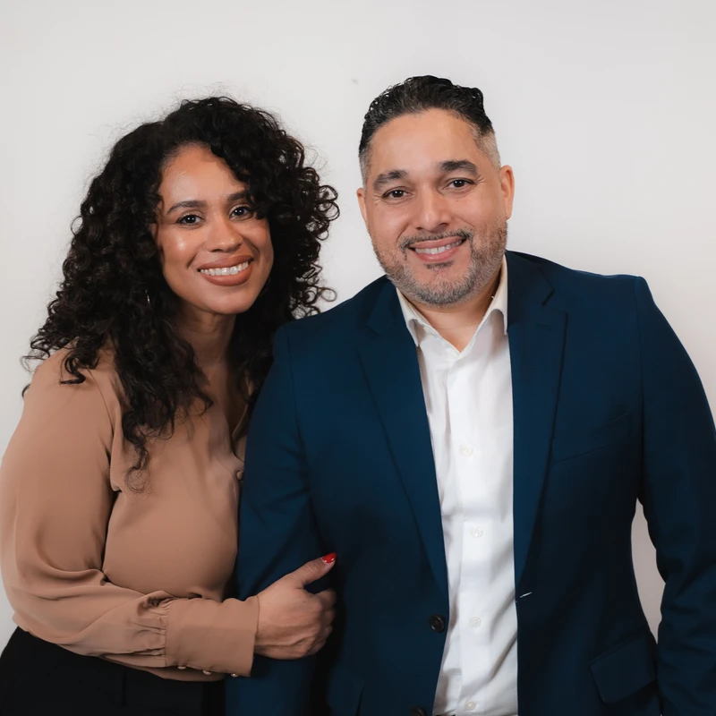 A professionally dressed man and woman smile while posing together against a plain white background. The woman has her arm linked with the man's.