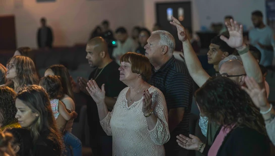 A diverse group of people of various ages stand together in a dimly lit room, many with their hands raised in prayer and worship during a service.
