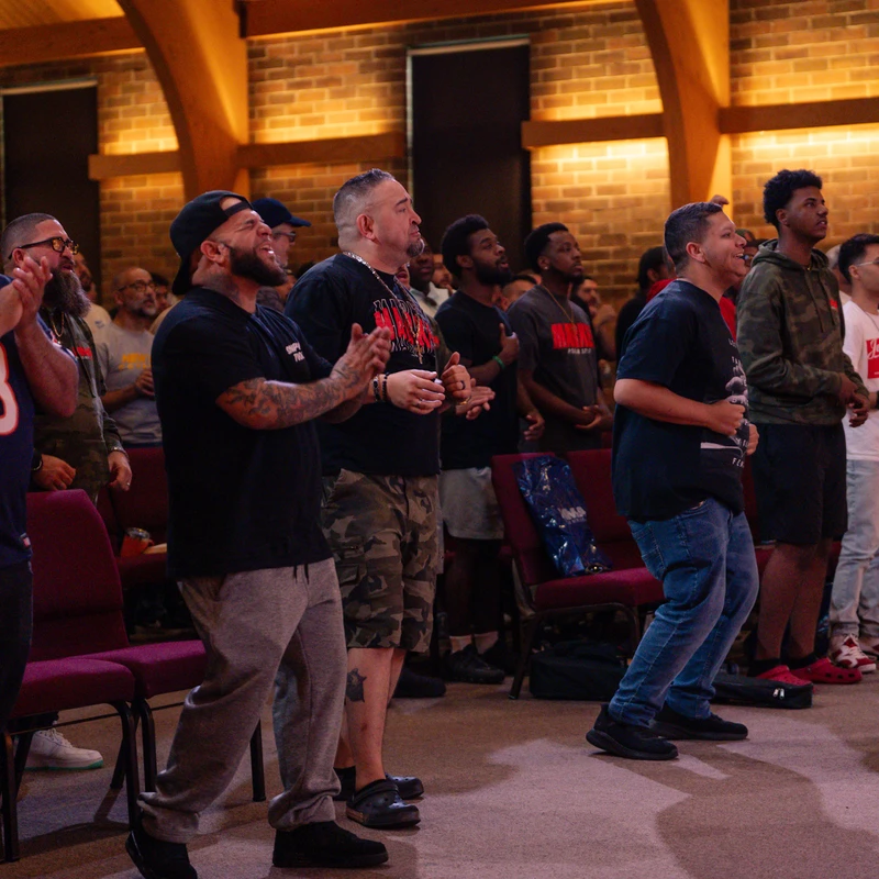 A diverse group of men standing in rows, clapping and singing with engaged expressions during a service in a warmly lit room with brick walls.