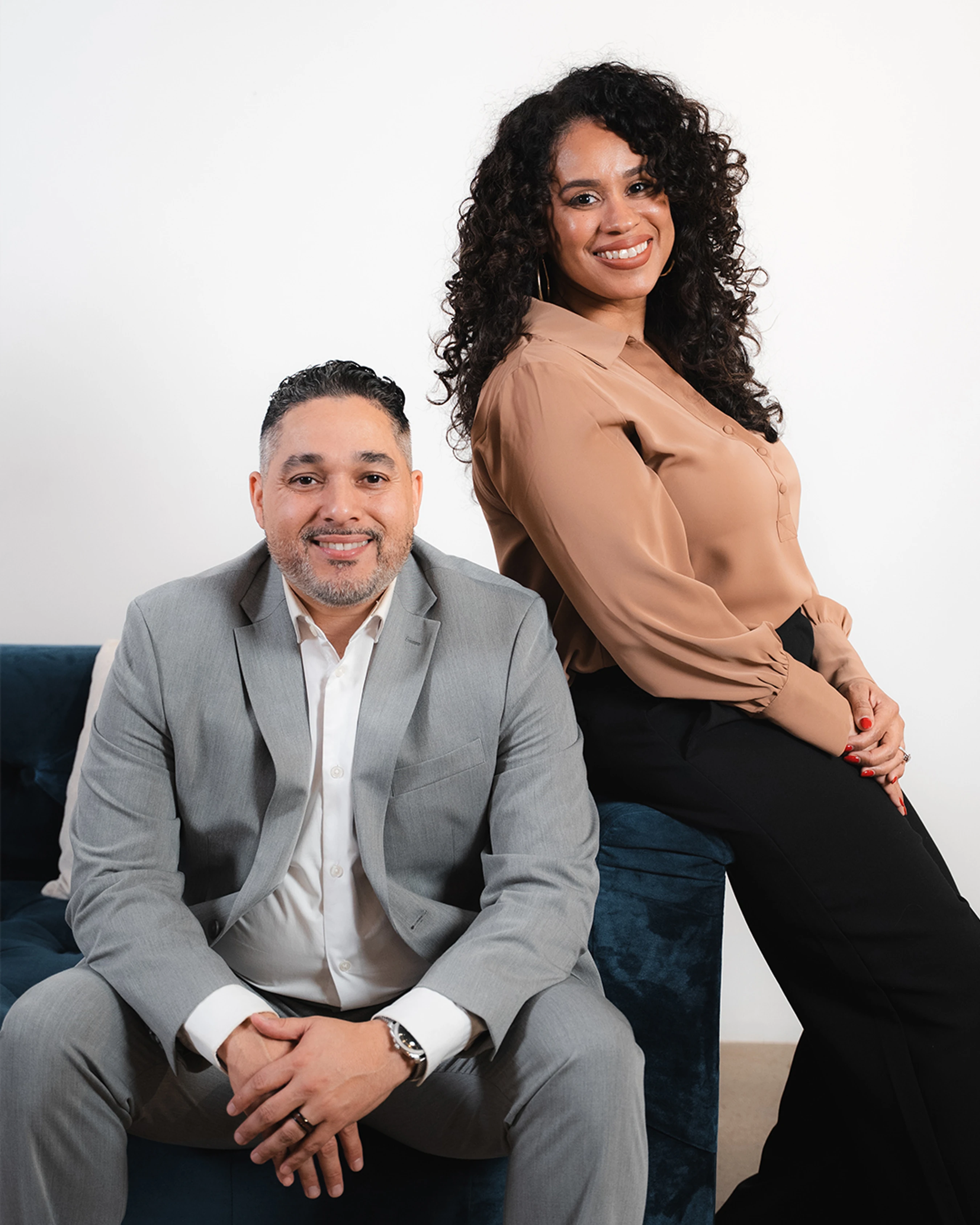 A man in a grey suit sits on a blue couch, smiling. A woman with curly hair in a tan blouse stands behind him, also smiling, in a studio portrait.