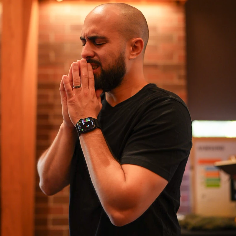 A man with a shaved head and beard stands indoors with his eyes closed and hands clasped in front of his face in a prayerful or emotional pose.