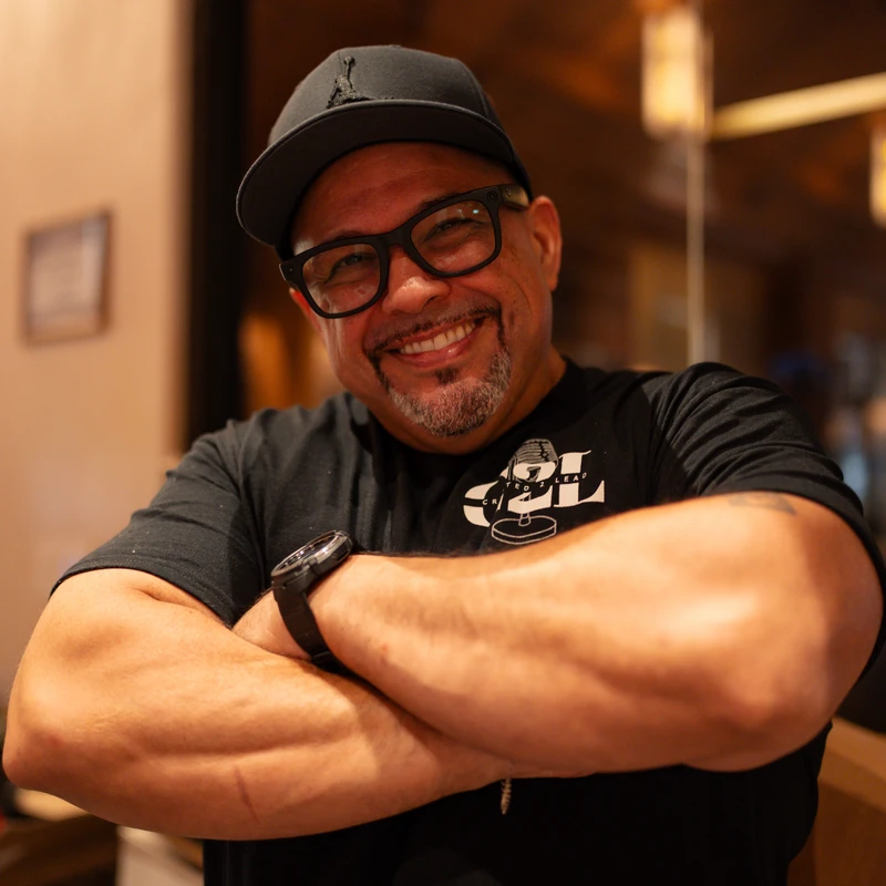 A medium shot of a smiling man wearing a black t-shirt, glasses, and a baseball cap, standing indoors with his muscular arms crossed.