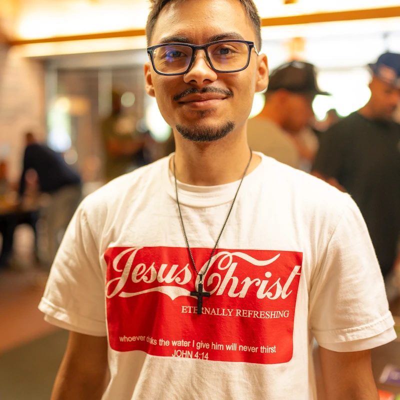 A young man with glasses and a goatee smiles while wearing a white t-shirt with the words 'Jesus Christ, Eternally Refreshing' in red.